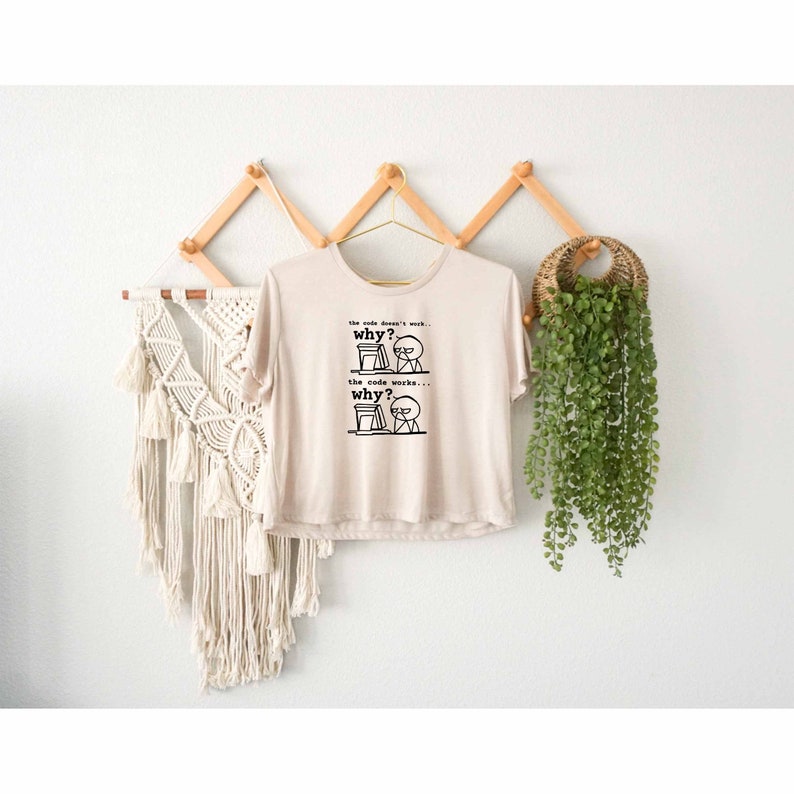 Puede incluir: Una camiseta beige con un gr&aacute;fico negro de una figura de palo frente a una computadora. El texto en la camiseta dice "the code doesn't work... why? the code works... why?"