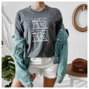 以下が含まれることがあります： コンピューターの前に立つ棒人間の白いグラフィックプリントが施されたダークグレーのロングスリーブシャツ。シャツのテキストは「the code doesn't work... why?」と「the code works... why?」です