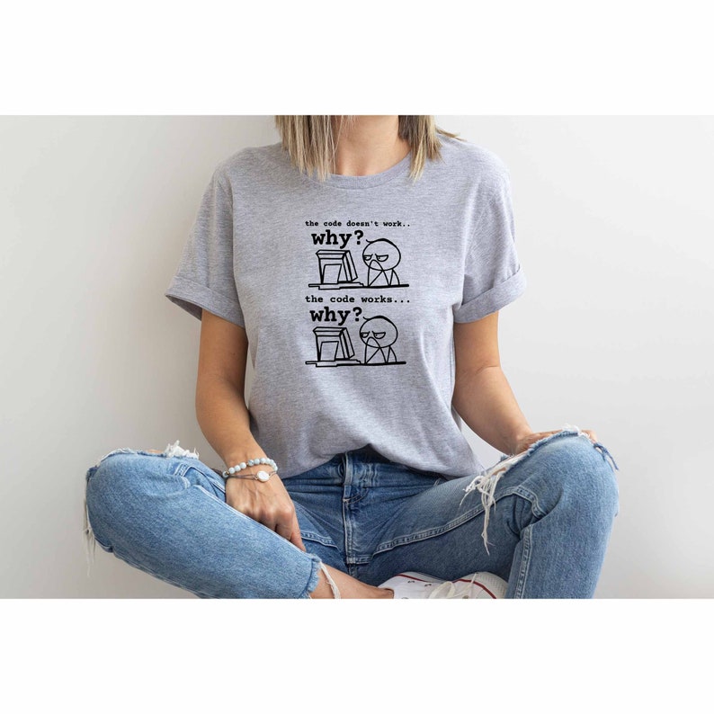 以下が含まれることがあります： コンピューターの前に座っている人の白黒のイラストが描かれたグレーのTシャツ。シャツのテキストは「the code doesn't work... why?」と「the code works... why?」です