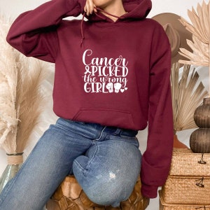 Puede incluir: Una sudadera con capucha color burdeos con un gráfico blanco que dice "Cancer & picked the wrong girl" con una cinta rosa y guantes de boxeo.