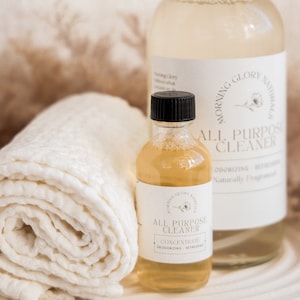 May include: Two clear glass bottles of all-purpose cleaner with white labels and black text. The labels read "Morning Glory Naturals" and "All Purpose Cleaner". A white rolled-up cloth and a smaller bottle are in front.