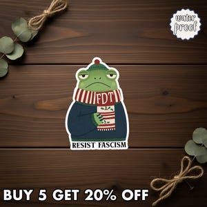 May include: A waterproof sticker featuring a grumpy green frog wearing a blue sweater, a red and white striped scarf with "FDT" on it, and a green beanie. The sticker also has the words "RESIST FASCISM" at the bottom.