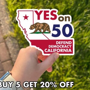 May include: A sticker in the shape of California with the text "YES on 50" and "DEFEND DEMOCRACY CALIFORNIA." The sticker features a brown bear and a red star. The background is green grass.