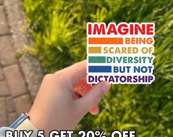 May include: A rectangular sticker with the text "IMAGINE BEING SCARED OF DIVERSITY BUT NOT DICTATORSHIP" in a colorful, retro-style design. The sticker is held against a background of green grass.