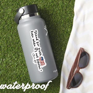 May include: A gray insulated water bottle with a black lid and handle. A sticker with the text "Stand for truth. Even when it's not popular." and an American flag is on the bottle. Wooden sunglasses rest on a white towel.