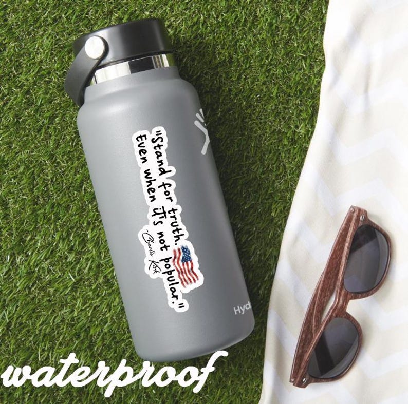 May include: A gray insulated water bottle with a black lid and handle. A sticker with the text "Stand for truth. Even when it's not popular." and an American flag is on the bottle. Wooden sunglasses rest on a white towel.