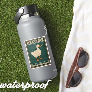 May include: A gray water bottle with a black lid and a sticker that reads "BECOME UNGOVERNABLE" featuring a goose with a knife. The word "waterproof" is printed at the bottom. Wooden-framed sunglasses rest on a white cloth.