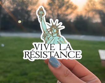 May include: A sticker featuring the Statue of Liberty with the words "VIVE LA RÉSISTANCE" in black text. The Statue of Liberty is depicted in shades of green and gold, holding a torch. The sticker is held by a hand with blue nail polish.