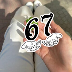 May include: A hand holding a green leaf with the numbers "67" in black. Below the numbers are two illustrations of hands with open palms. The person is wearing white pants and white shoes.