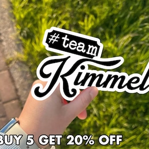 May include: A white sticker with the text "#team Kimmel" in a black, stylized font. The sticker is held against a background of green grass. The text "BUY 5 GET 20% OFF" is at the bottom.