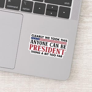 May include: A rectangular sticker with the text "CLEARLY WE TOOK THIS ANYONE CAN BE PRESIDENT THING A BIT TOO FAR" in red, white, and blue. The sticker is on a gray laptop.