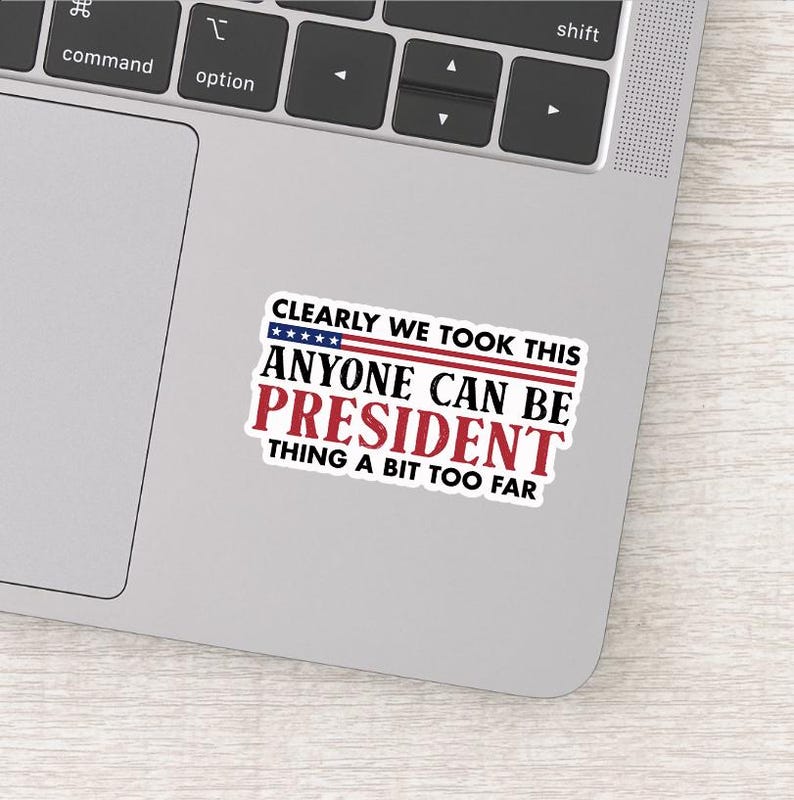 May include: A rectangular sticker with the text "CLEARLY WE TOOK THIS ANYONE CAN BE PRESIDENT THING A BIT TOO FAR" in red, white, and blue. The sticker is on a gray laptop.