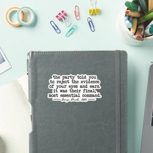 May include: A gray notebook with the quote "the party told you to reject the evidence of your eyes and ears. It was their final, most essential command. George Orwell, 1984." Colorful office supplies surround the notebook.