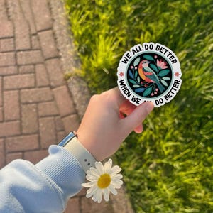 May include: A round sticker with the text "WE ALL DO BETTER WHEN WE DO BETTER" encircling a colorful illustration of a bird, flowers, and leaves. The sticker is held in a hand, with a daisy and a watch visible.