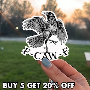 May include: A black and white sticker featuring a detailed illustration of a crow with outstretched wings and the text "F-CAW-F". The sticker is held up against a blurred outdoor background.