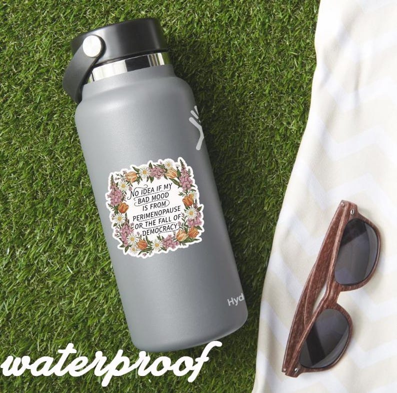 May include: A gray insulated water bottle with a sticker that reads "No idea if my bad mood is from perimenopause or the fall of democracy." The sticker is surrounded by a floral design. Wooden sunglasses rest on a white towel.