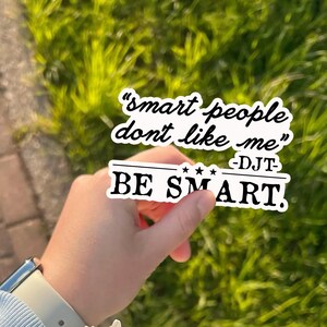 May include: A white sticker with black text that reads "smart people don't like me" and "BE SMART." The sticker is held in front of a blurred green background.