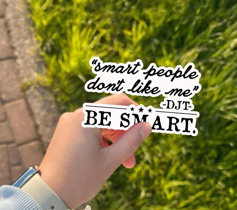 May include: A white sticker with black text that reads "smart people don't like me" and "BE SMART." The sticker is held in front of a blurred green background.