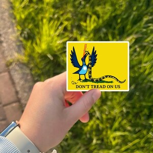 May include: A yellow sticker featuring a blue bird with black wings and a yellow and black snake. The sticker has the text "DON'T TREAD ON US" in black. The sticker is held in front of a green grassy background.