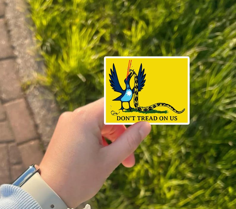 May include: A yellow sticker featuring a blue bird with black wings and a yellow and black snake. The sticker has the text "DON'T TREAD ON US" in black. The sticker is held in front of a green grassy background.