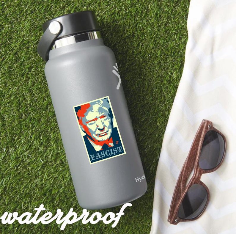 May include: A gray Hydro Flask water bottle with a sticker featuring a portrait with the word "Fascist". The image also includes a pair of wooden-framed sunglasses and the word "waterproof". The items are on a green grassy surface.