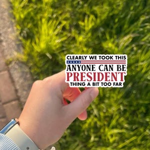 May include: A rectangular sticker with the text "CLEARLY WE TOOK THIS ANYONE CAN BE PRESIDENT THING A BIT TOO FAR." The sticker has a white background with red and blue text and a blue and white stars and stripes design.