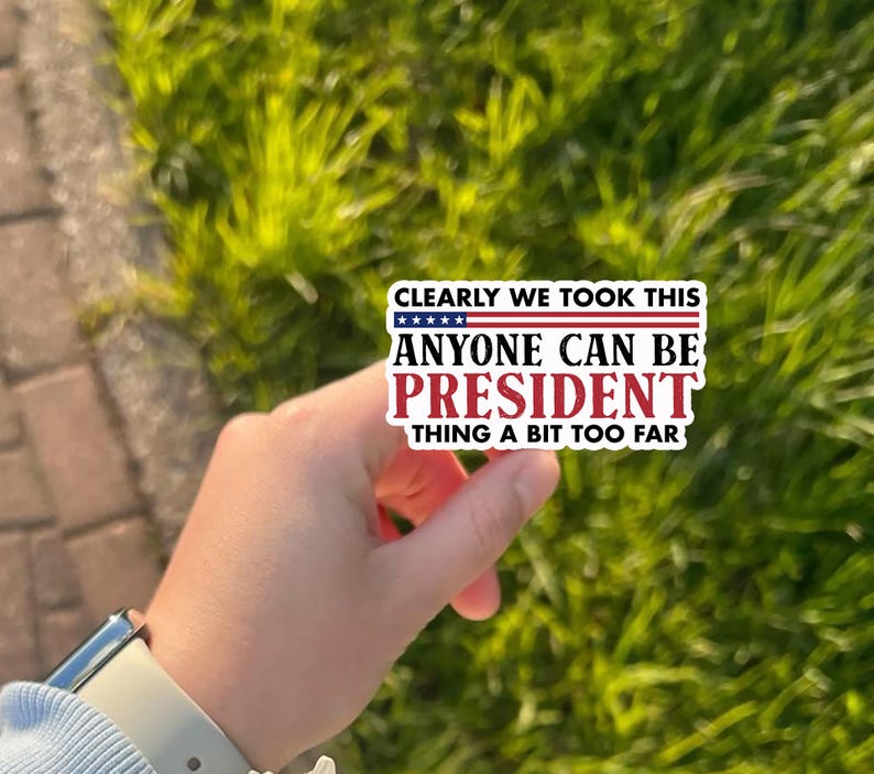 May include: A rectangular sticker with the text "CLEARLY WE TOOK THIS ANYONE CAN BE PRESIDENT THING A BIT TOO FAR." The sticker has a white background with red and blue text and a blue and white stars and stripes design.