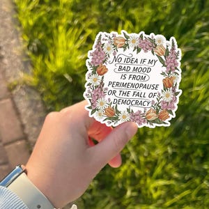 May include: A white sticker with a floral border featuring the text: "No idea if my bad mood is from perimenopause or the fall of democracy." The sticker has a variety of flowers in shades of pink, orange, and white.
