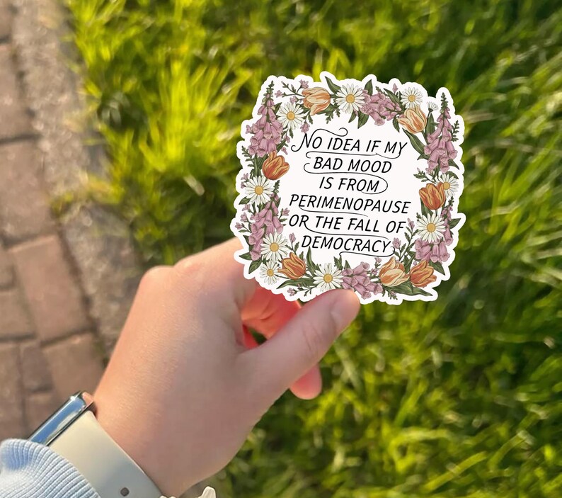 May include: A white sticker with a floral border featuring the text: "No idea if my bad mood is from perimenopause or the fall of democracy." The sticker has a variety of flowers in shades of pink, orange, and white.