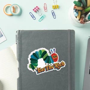 May include: A colorful sticker featuring a cartoon caterpillar with a red face and blue antennae. The caterpillar is surrounded by green and blue segments. The sticker has the text "Eat The Rich" in a stylized font. The sticker is on a gray notebook.
