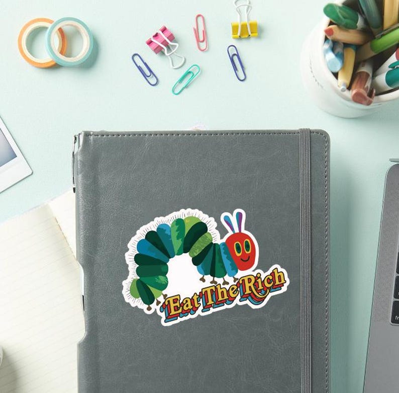 May include: A colorful sticker featuring a cartoon caterpillar with a red face and blue antennae. The caterpillar is surrounded by green and blue segments. The sticker has the text "Eat The Rich" in a stylized font. The sticker is on a gray notebook.