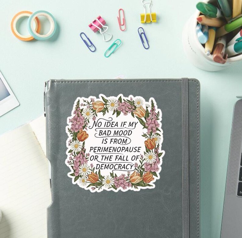 May include: A sticker with the text "No idea if my bad mood is from perimenopause or the fall of democracy" surrounded by a floral wreath. The sticker is on a gray notebook, with office supplies in the background.