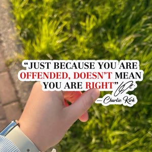 May include: A white sticker with the quote "JUST BECAUSE YOU ARE OFFENDED, DOESN'T MEAN YOU ARE RIGHT" by Charlie Kirk. The sticker is held in front of a background of green grass and a brick walkway.