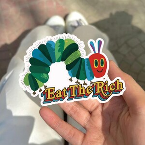 May include: A colorful sticker featuring a cartoon caterpillar with a red face, blue and green body segments, and the text "Eat The Rich" in a retro font. The sticker is held in a hand against a blurred background.