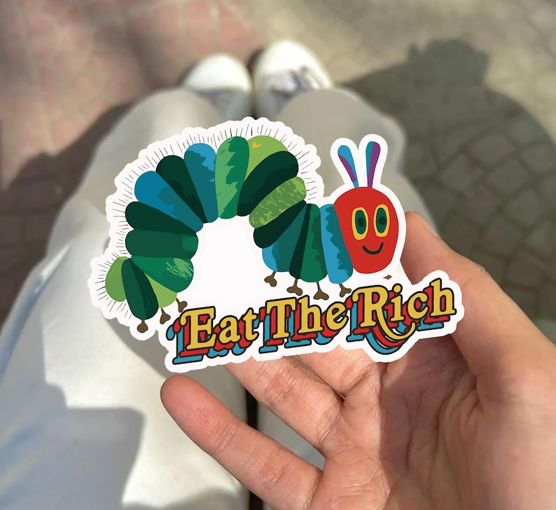 May include: A colorful sticker featuring a cartoon caterpillar with a red face, blue and green body segments, and the text "Eat The Rich" in a retro font. The sticker is held in a hand against a blurred background.