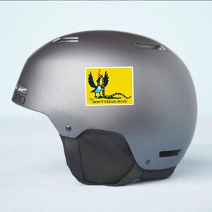 May include: A gray helmet with a sticker featuring a yellow rectangle with a coiled snake and the words "DON'T TREAD ON US". The sticker has a blue bird-like creature with a snake in its beak.