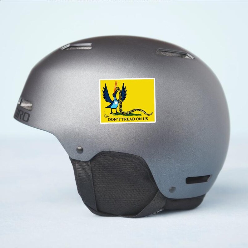 May include: A gray helmet with a sticker featuring a yellow rectangle with a coiled snake and the words "DON'T TREAD ON US". The sticker has a blue bird-like creature with a snake in its beak.