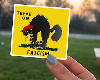 May include: A yellow sticker with a black cat wearing a red hat, biting a yellow and black snake. The sticker reads "TREAD ON FASCISM".
