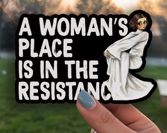 May include: A black sticker with the text "A Woman's Place Is In The Resistance" in white. The sticker features a cartoon illustration of a woman in a white gown. The background is blurred.