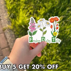 May include: A sticker featuring illustrations of a foxglove, daisy, and tulip. The flowers are in full bloom with green stems and leaves. The sticker has the words "Foxglove", "Daisy", and "Tulip" in green block letters. The background is blurred green grass.