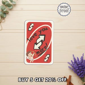 May include: A red and white sticker with the text "love you more!" and an illustration of Cupid. The sticker is waterproof and has a reverse card design. The sticker is on a wooden surface.