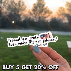 May include: A white sticker with the text "Stand for truth. Even when it's not popular." and an American flag graphic. The sticker is held up against a blurred outdoor background.