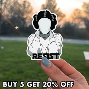 May include: A white sticker featuring a faceless illustration of a character with a distinctive hairstyle and a white outfit. The word "RESIST" is printed in black. The sticker is held by a hand with blue nail polish.