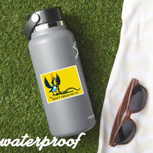 May include: A gray water bottle with a black lid and a sticker featuring a yellow flag with a coiled snake and the text "DON'T TREAD ON US." The word "waterproof" is written in white script. Wooden-framed sunglasses rest on a white towel.
