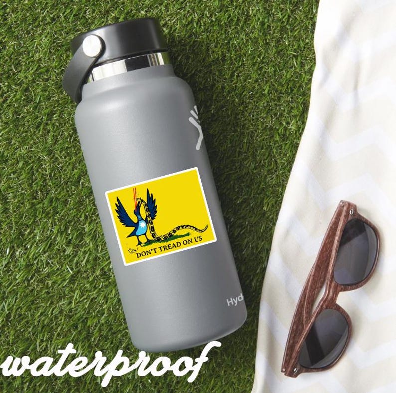 May include: A gray water bottle with a black lid and a sticker featuring a yellow flag with a coiled snake and the text "DON'T TREAD ON US." The word "waterproof" is written in white script. Wooden-framed sunglasses rest on a white towel.