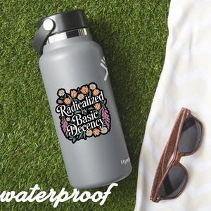 May include: A gray water bottle with a black lid and a sticker that reads "Radicalized by Basic Decency" surrounded by floral illustrations. The word "waterproof" is written at the bottom. Wooden-framed sunglasses rest on a white cloth.