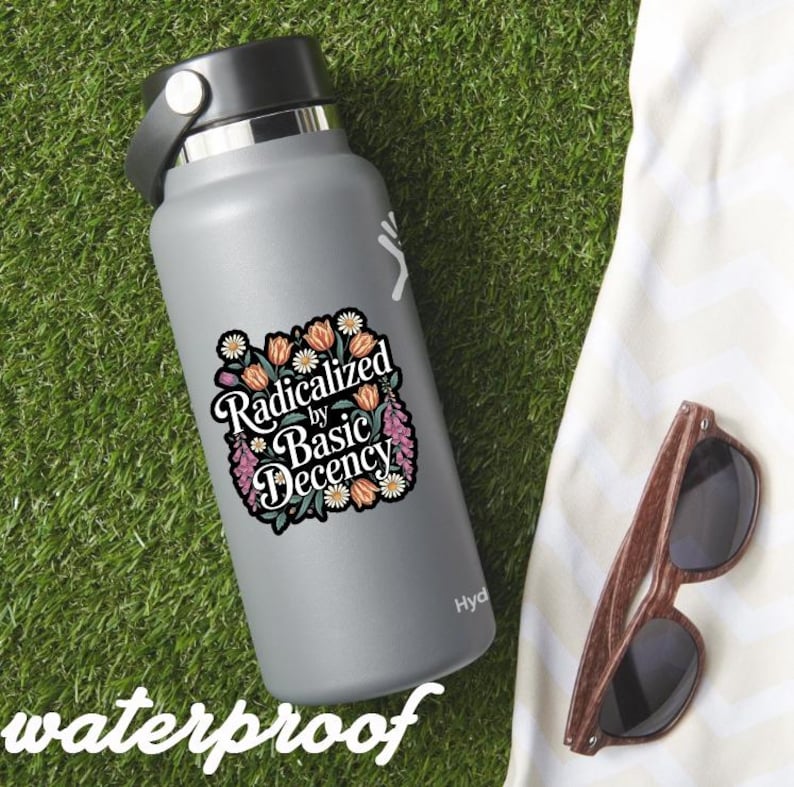 May include: A gray water bottle with a black lid and a sticker that reads "Radicalized by Basic Decency" surrounded by floral illustrations. The word "waterproof" is written at the bottom. Wooden-framed sunglasses rest on a white cloth.