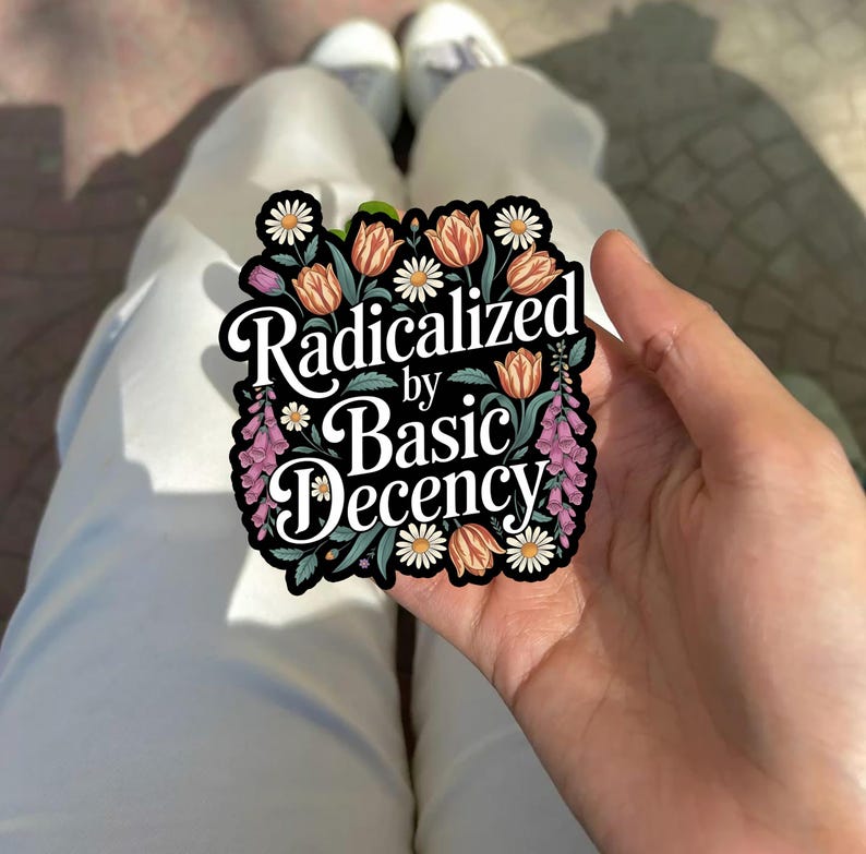 May include: A sticker with the text "Radicalized by Basic Decency" in a decorative font, surrounded by a floral border of daisies, tulips, and other flowers. The sticker is black with white, orange, pink, and green accents.