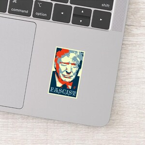 May include: A sticker featuring a stylized portrait with red, blue, and yellow tones. The word "FASCIST" is printed in a blue box at the bottom. The sticker is placed on a laptop.