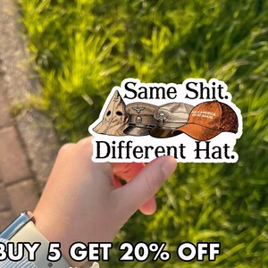 May include: A sticker with the text "Same Shit. Different Hat." features illustrations of a KKK hood, a military cap, and an orange baseball cap with "MAKE AMERICA GREAT AGAIN" printed on it.
